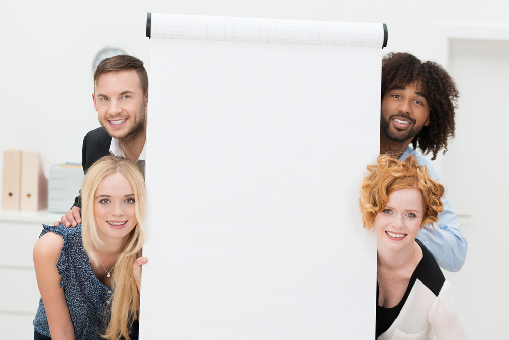 Smiling successful multiethnic business team with a blank white sheet of paper on a flip chart peering playfully around the sides at the camera